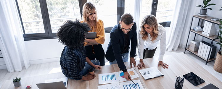 Four colleagues stand around a table reviewing printed charts and reports together