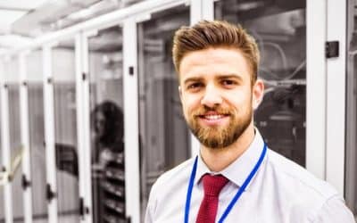 IT professional smiling in a server room while a colleague works on a laptop