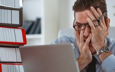 Frustrated employee with head in hands at a laptop beside stacked binders